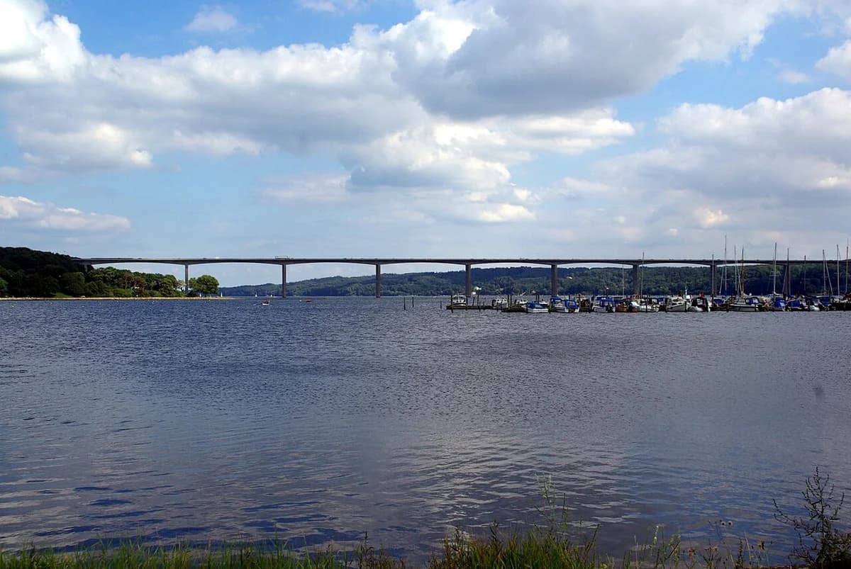 A long bridge crossing a fjord with boats and cloudy sky.