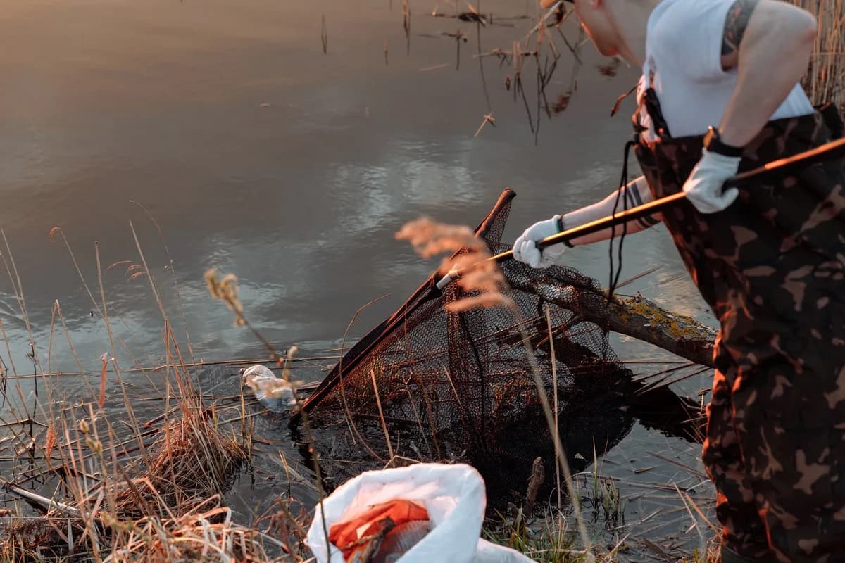 An adult in gloves and waders uses a large net to remove debris from the edge of a pond.